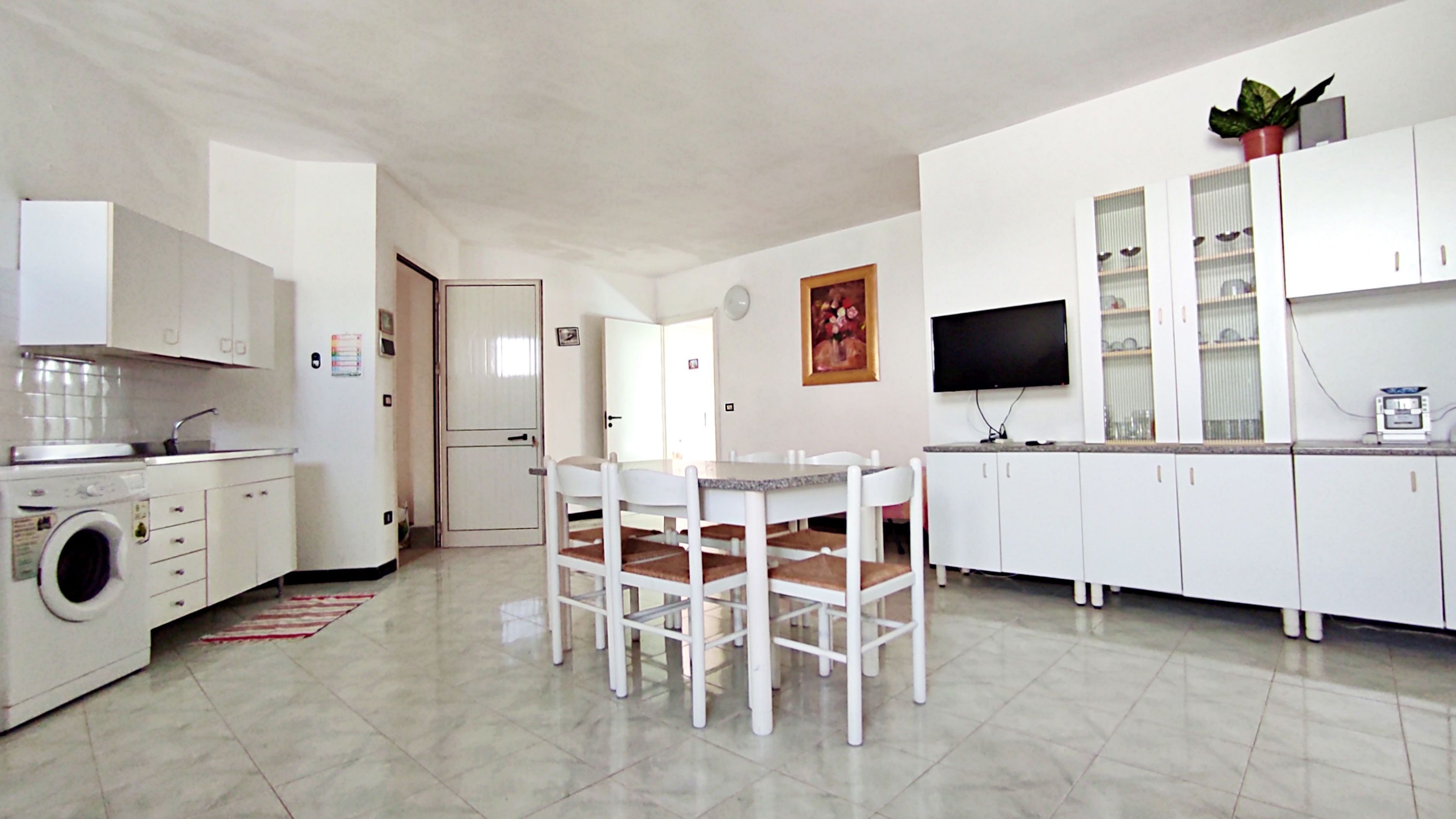 A compact kitchen with white cabinets, a washing machine, a dining table with chairs, a TV, and a potted plant.