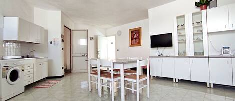 A compact kitchen with white cabinets, a washing machine, a dining table with chairs, a TV, and a potted plant.