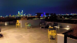 An outdoor seating area with chairs and tables, a tiled floor, and a city skyline in the background.
