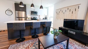 A modern kitchen with a central island, bar stools, a flat-screen TV, and a decorative macrame wall hanging.