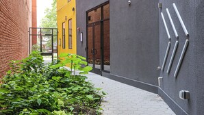 A narrow alleyway with a brick wall on one side and a modern building with a gray facade and a yellow wall on the other. There are metal handles on the gray wall and a small garden area with green plants.