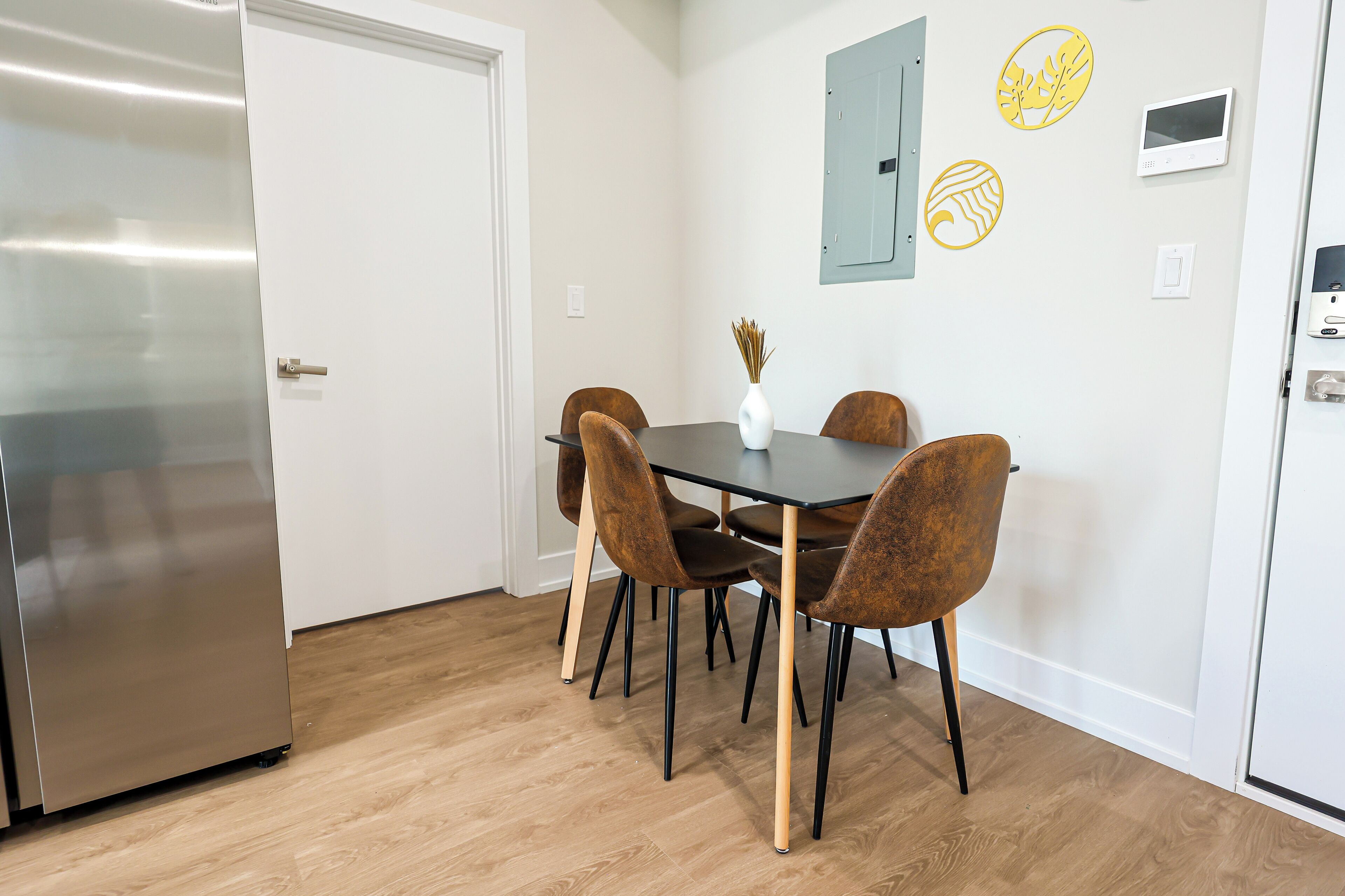 A modern kitchen with a stainless steel refrigerator, a wooden dining table with four chairs, and a vase with dried plants on the wall.
