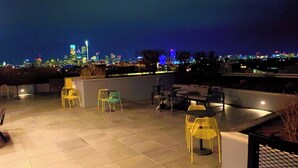 Un espacio al aire libre con sillas y mesas, un horizonte urbano al fondo y vista de la ciudad de noche.