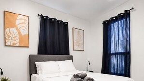 Una cama bien hecha con sábanas blancas, cabecera gris y toallas enrolladas sobre la cama. Hay un tendedero de metal negro y una mesita con una planta y una lámpara.