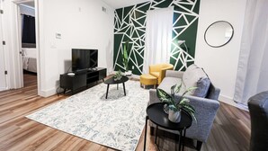 Una sala de estar moderna con sofá, sillón y mesa de centro. Hay un televisor en un mueble bajo y una planta en una mesita.