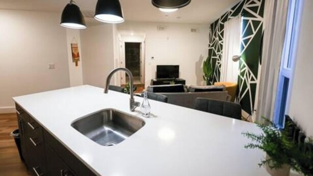 Cocina moderna con una isla central, lámparas colgantes negras y vistas a una sala de estar con sofá y televisor.