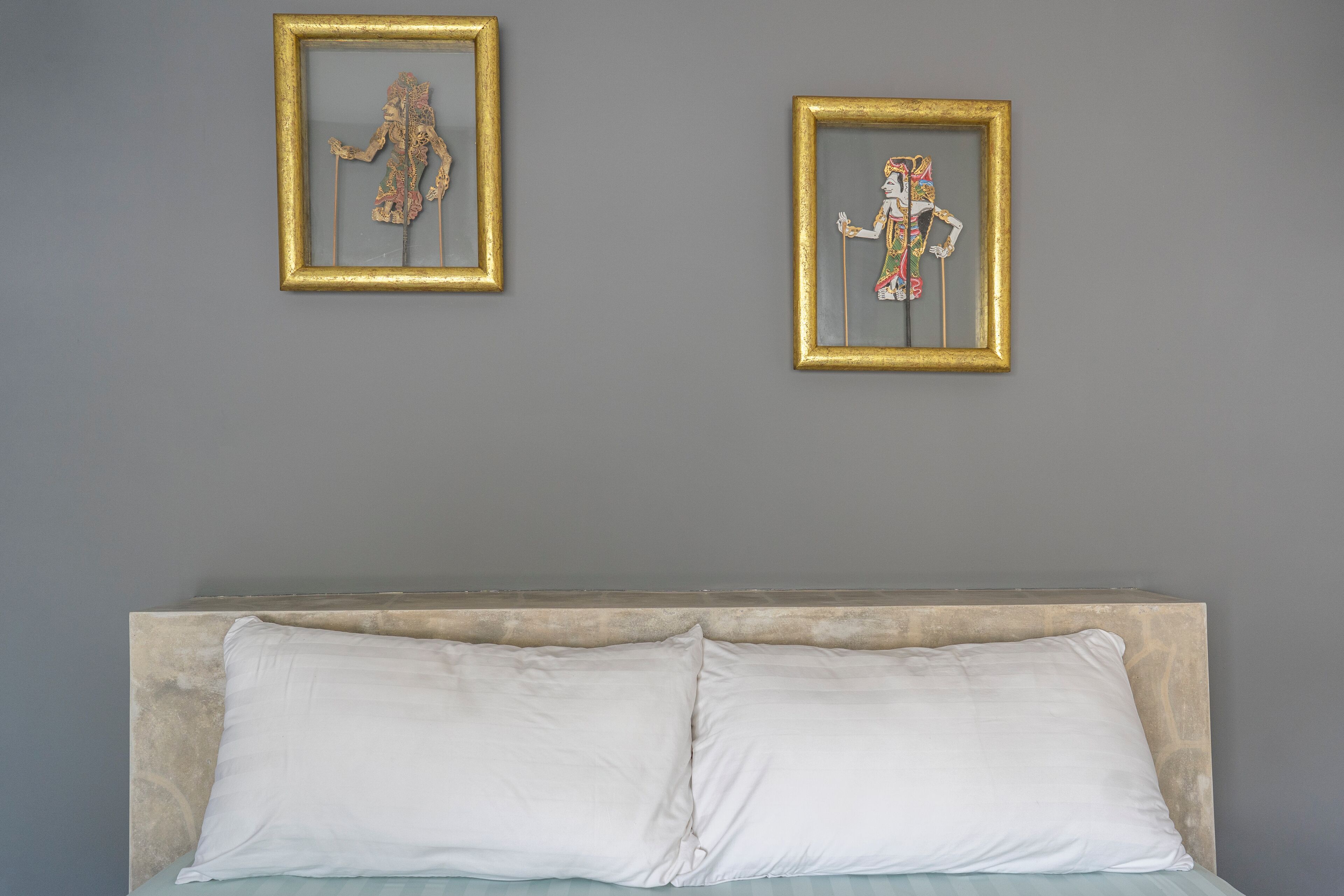 Un lit avec des oreillers blancs et une tête de lit, deux œuvres d’art encadrées accrochées au mur, et un mur uni de couleur grise.