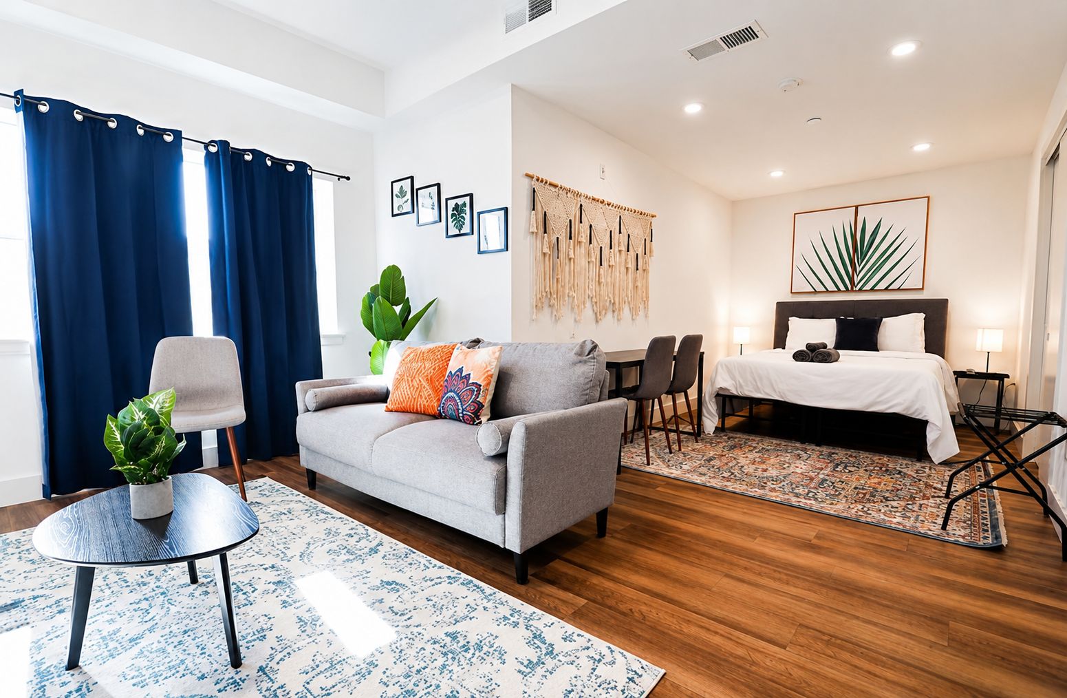 Une chambre d’hôtel moderne avec un canapé gris, une table à manger avec des chaises, un lit avec une couette blanche et une grande plante.