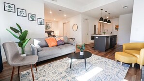Una sala de estar moderna con un sofá gris, una mesa redonda y un sillón amarillo. Se observa una zona de cocina con una isla gris e iluminación colgante.