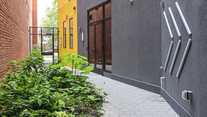 Un callejón estrecho con un edificio amarillo, otro gris y una pared de ladrillo. Hay plantas en primer plano y un camino con baldosas hexagonales.