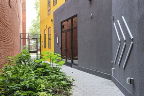 Un vialetto stretto con un edificio giallo, uno grigio e un muro in mattoni. In primo piano ci sono delle piante e un percorso con piastrelle esagonali.