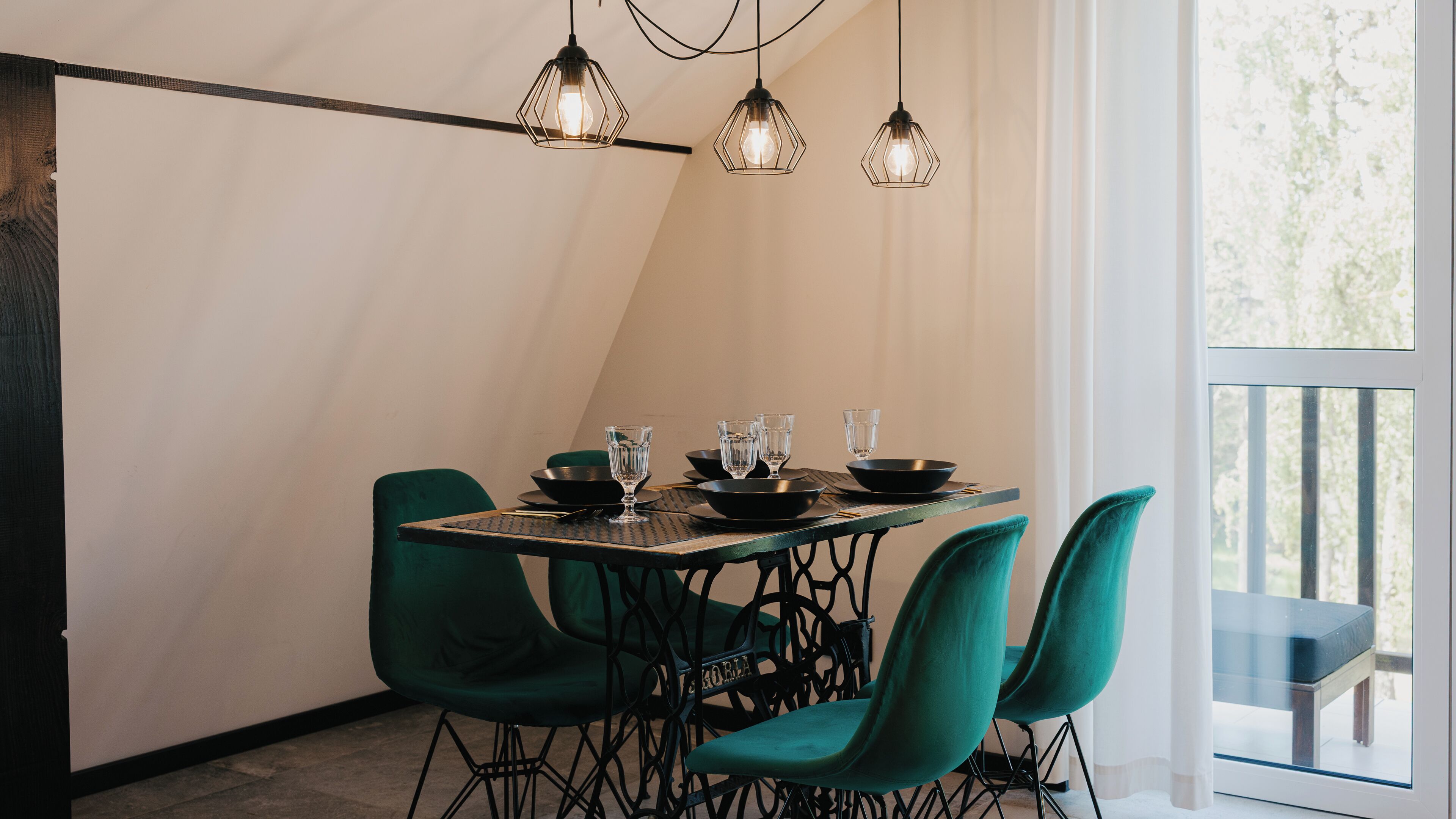 Un coin repas avec une table dressée pour quatre personnes, des chaises à l’assise rembourrée de tissu vert et des suspensions avec des ampoules apparentes.