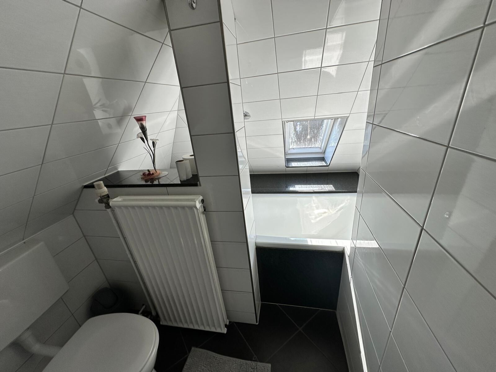 A compact bathroom with white tiles, a toilet, a radiator, and a skylight.