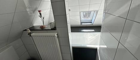 A compact bathroom with white tiles, a toilet, a radiator, and a skylight.