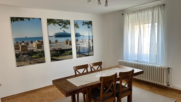 A dining area with a wooden table and chairs, three framed pictures on the wall, a window with sheer curtains, and a radiator. - Large apartment in the center of Plön within walking distance to the lake/shops. Children's room (Plön)