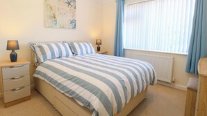A bedroom with a bed, two bedside tables, a nightstand, a radiator, and a window with curtains. - Bankside Cottage (King's Lynn)