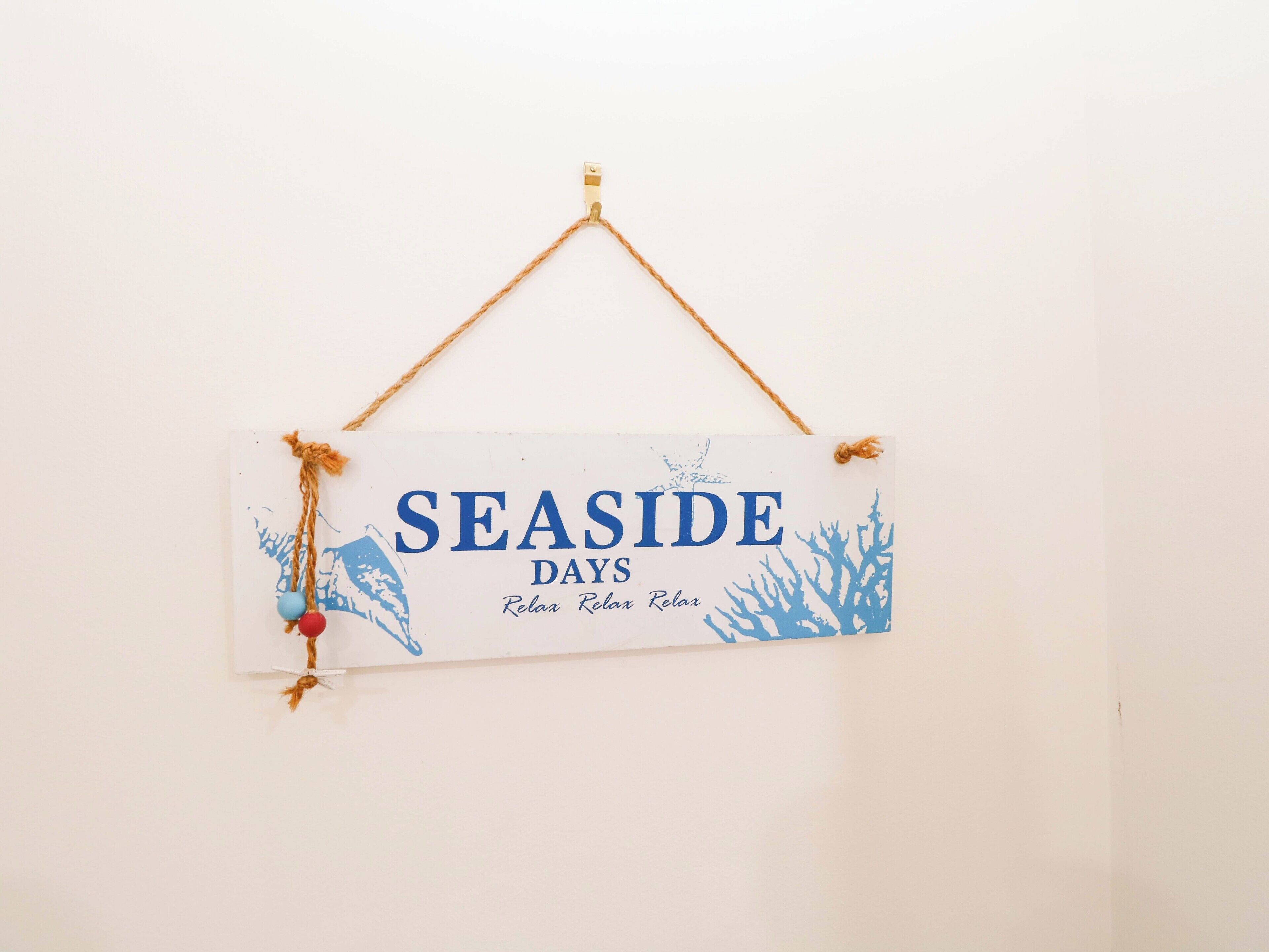 A sign with the text "SEASIDE DAYS" and "Relax Relax Relax" on a white background, hanging on a wall with a simple rope.