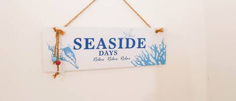 A sign with the text "SEASIDE DAYS" and "Relax Relax Relax" on a white background, hanging on a wall with a simple rope.