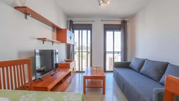 Ein modernes Wohnzimmer mit einer Couch, Fernseher, Holztisch und einem Balkon mit Vorhängen.