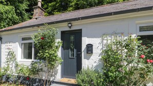 A white house with a dark door, a small porch, and a garden with various plants and flowers. - Nithbank Cottage (Thornhill)