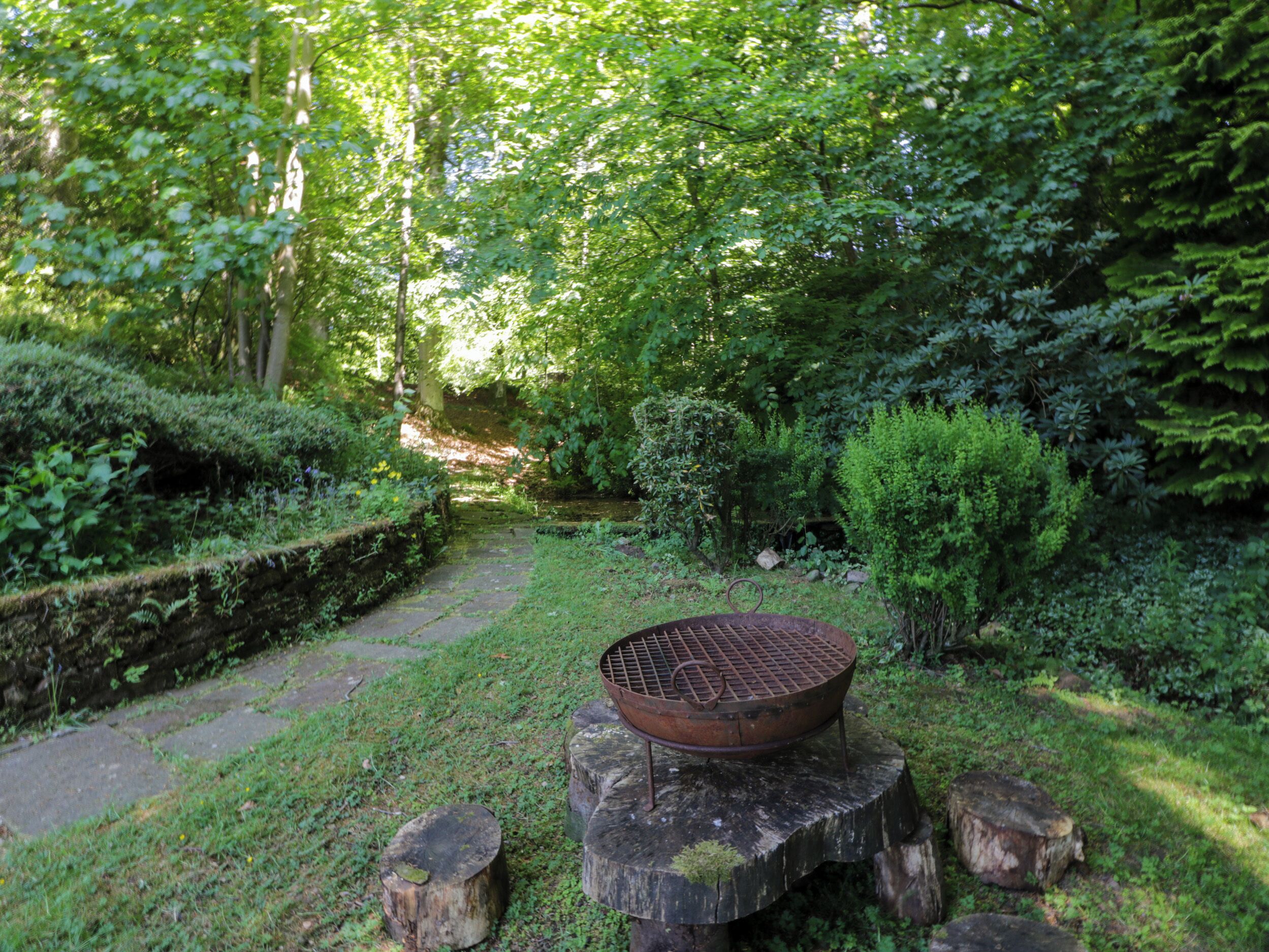 A rustic outdoor setting with a metal grill on a wooden stump, surrounded by lush greenery and a stone pathway.