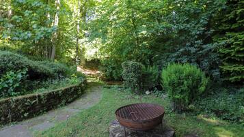 A rustic outdoor setting with a metal grill on a wooden stump, surrounded by lush greenery and a stone pathway.