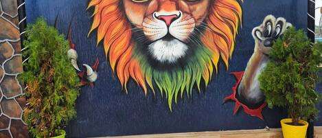 A mural of a lion wearing a cap with the logo "CITY X KITCHEN" on a blue wall. There are two potted plants in front of the mural.