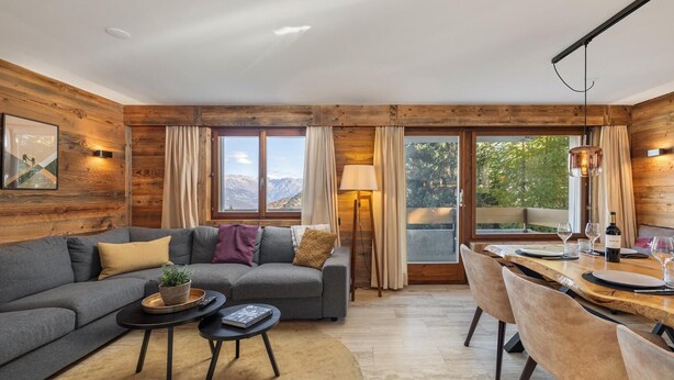 A modern living room with a grey sofa, a small round table, and a dining area with chairs. Large windows offer a view of mountains. - Apartment Diana 9 - Recently Renovated (Riddes)