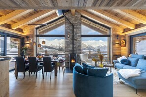 Un amplio salón con techo alto, vigas de madera, chimenea de piedra, ventanas grandes con vistas a la montaña y muebles modernos.