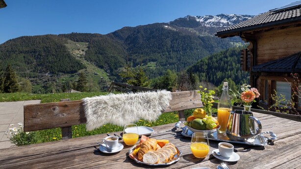 A wooden table set with breakfast items, a bottle, and a glass of orange juice, overlooking a mountainous landscape. - Chalet Belle Vue Alpine Charm With a View to Remember (Riddes)
