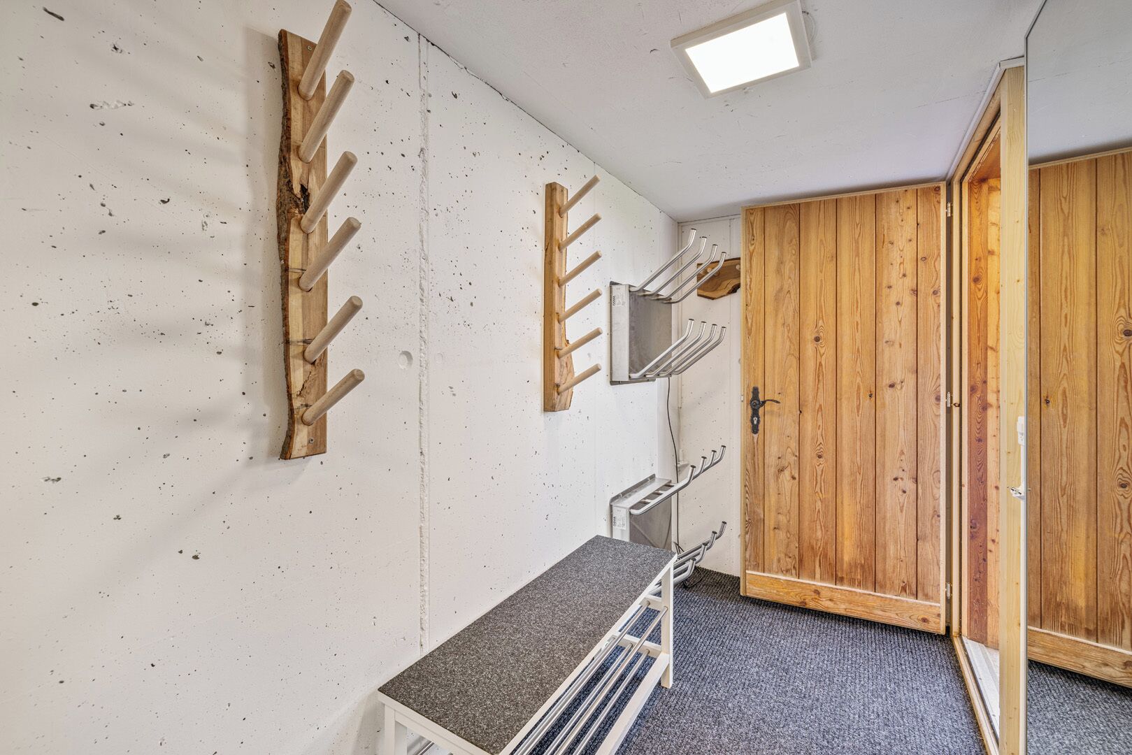 Ein Zimmer mit Holztür, einer weißen Wand mit Holzgestell, einem kleinen Tisch und einer Deckenleuchte.