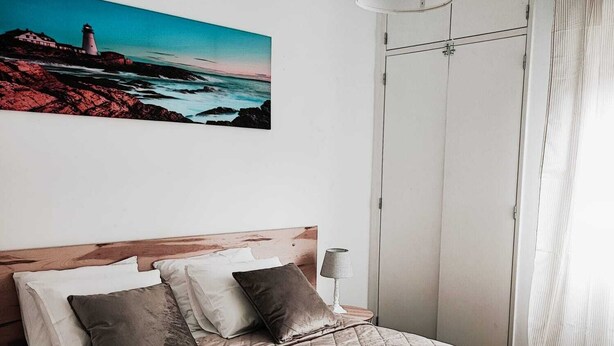 Een netjes opgemaakt bed met een gewatteerde spreideken, een houten hoofdbord en een bedlampje. Daarboven hangt een schilderij van een kustlandschap.