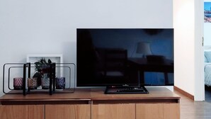 Een flatscreen televisie op een houten kast met een afstandsbediening, een glazen plank met decoratieve objecten en een witte muur.