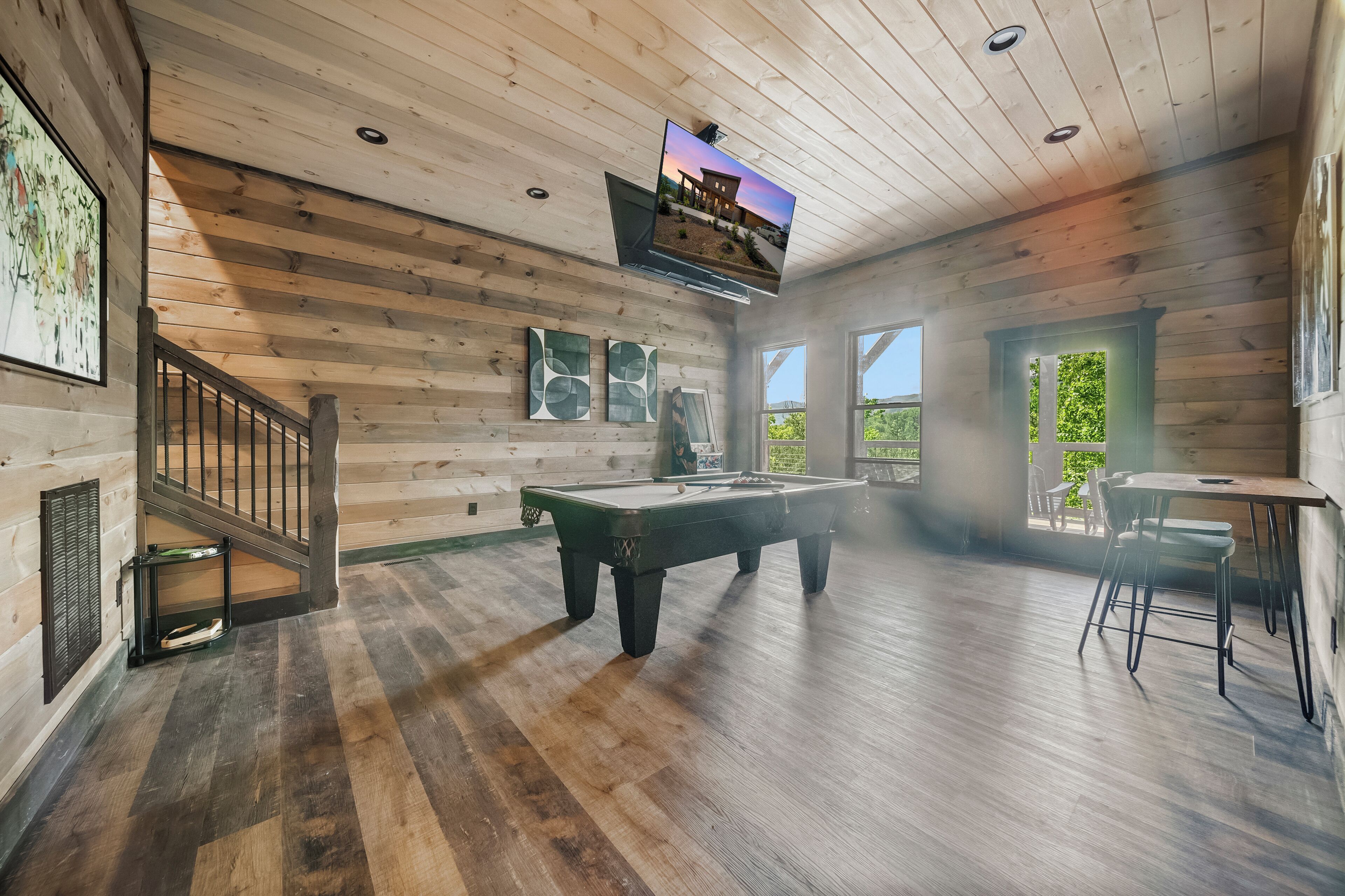 A wooden interior with a pool table, a TV mounted on the ceiling, and a staircase.