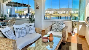 Un balcon ensoleillé, meublé de mobilier en osier, offrant une vue sur un paysage côtier, et une porte vitrée donnant sur l’intérieur.