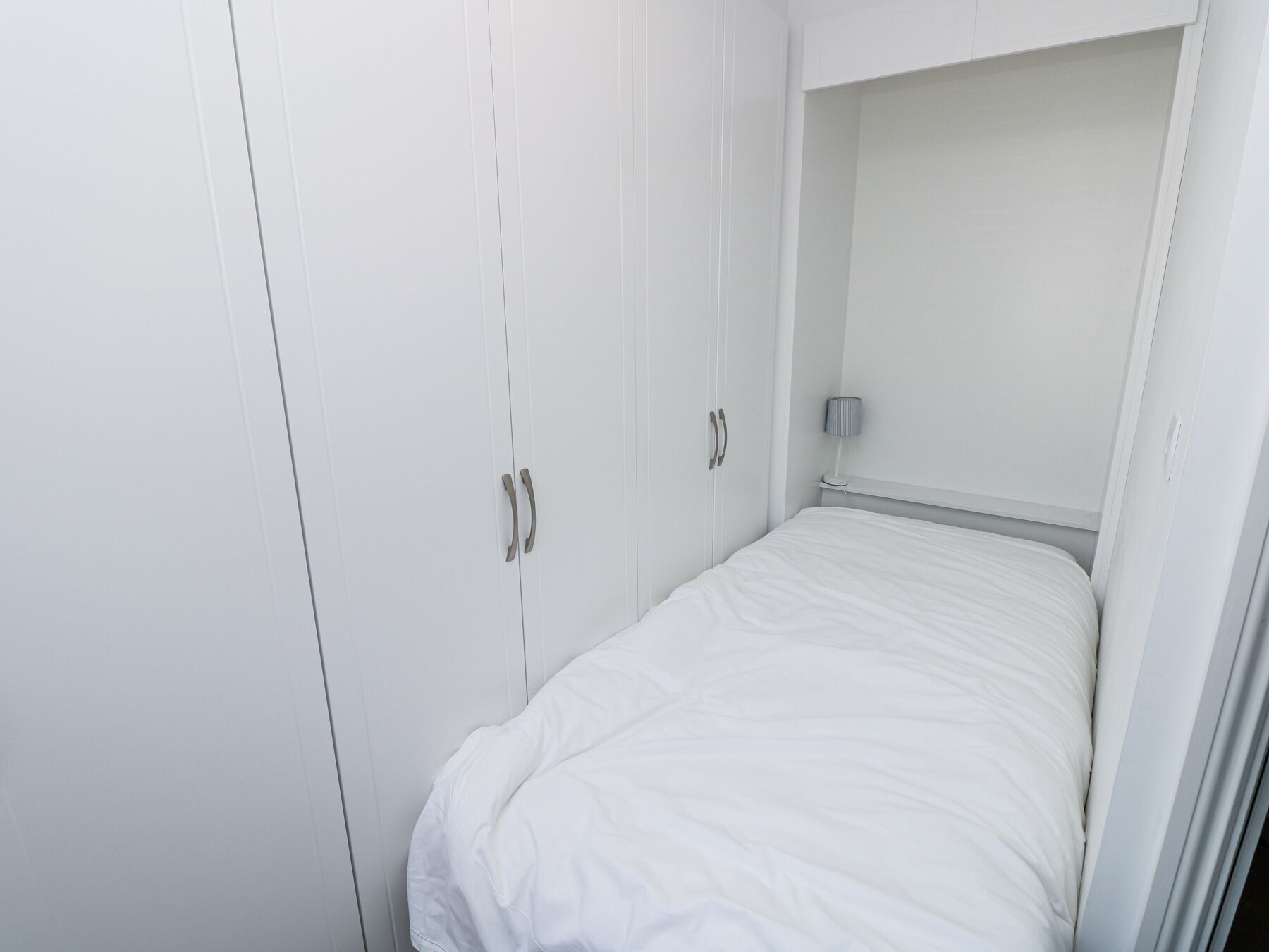 A white bedroom with a single bed, a wardrobe with two doors, and a bedside table with a lamp.
