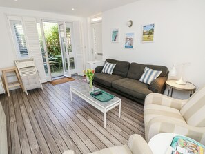 Uma sala de estar com piso de madeira, um sofá, uma mesa de centro, uma mesinha de cabeceira com abajur e uma porta que dá para uma varanda.