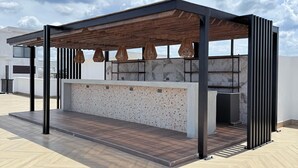 Un espace extérieur moderne, aménagé avec un coin salon, une pergola en bois, un comptoir en marbre et un sol en parquet disposé en chevrons.