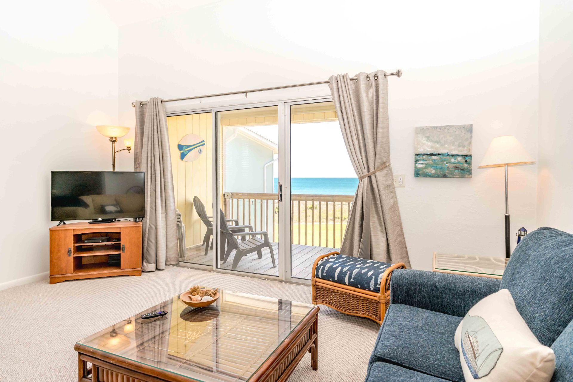 Ein Wohnzimmer mit Fernseher, einem Glastisch, einem Rattansofa und Blick auf den Strand durch eine Glasschiebetür.