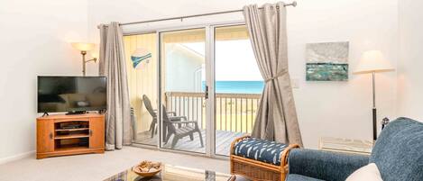 Ein Wohnzimmer mit Fernseher, einem Glastisch, einem Rattansofa und Blick auf den Strand durch eine Glasschiebetür.