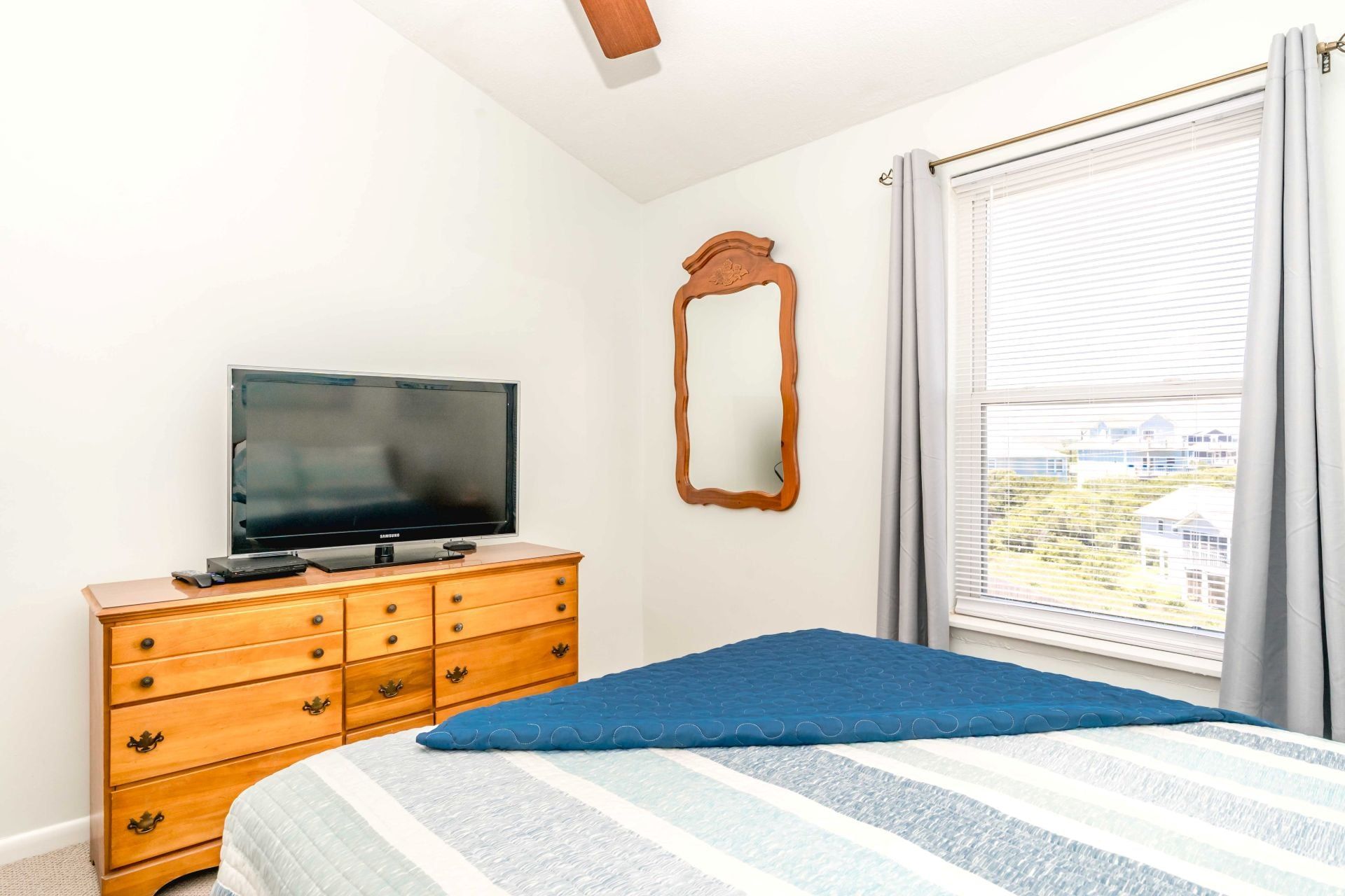 A bedroom with a bed, a dresser, a television, a mirror, and a window with blinds.