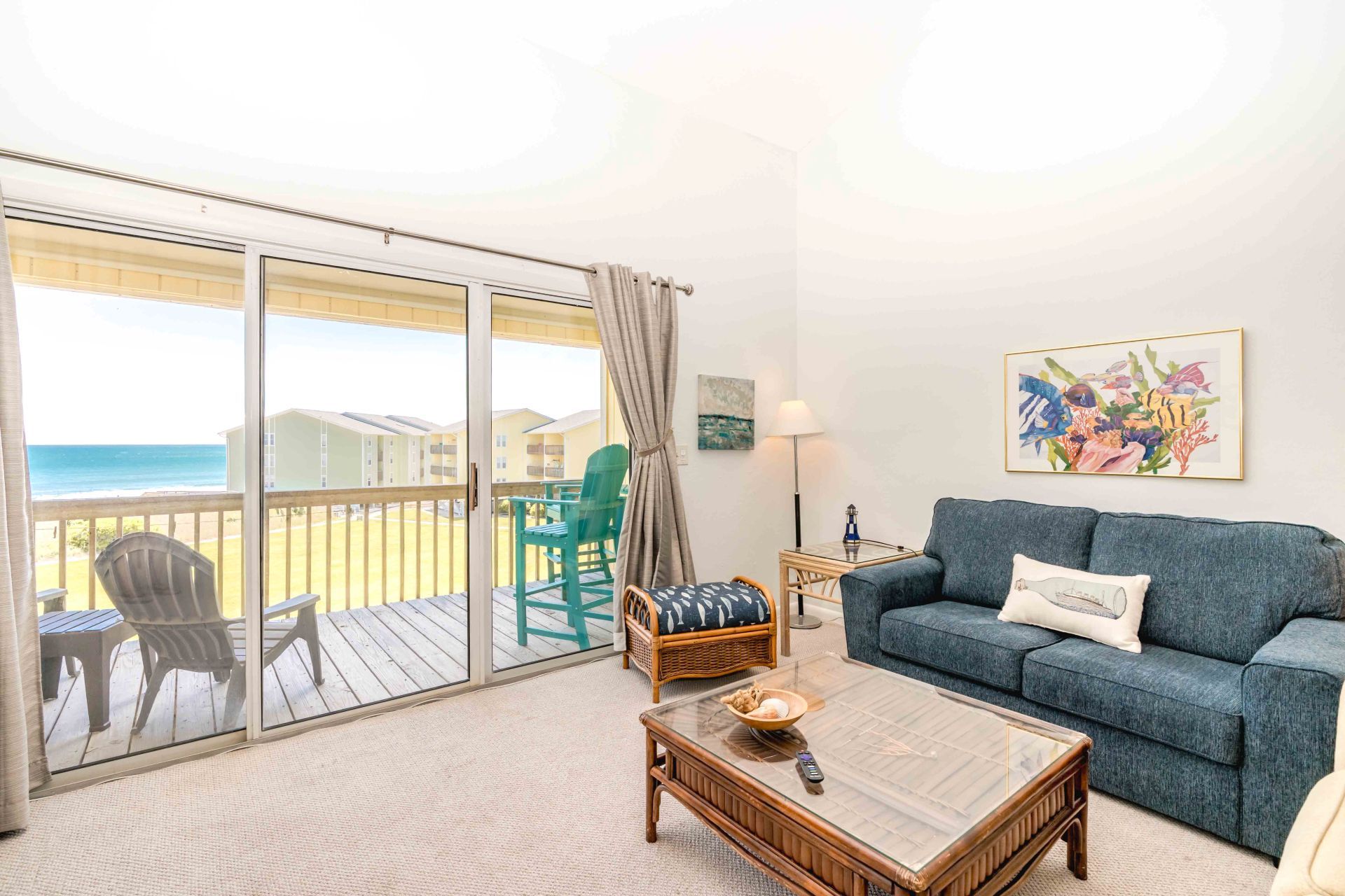 A living room with a glass coffee table, a blue sofa, and a view of the ocean through a sliding glass door.