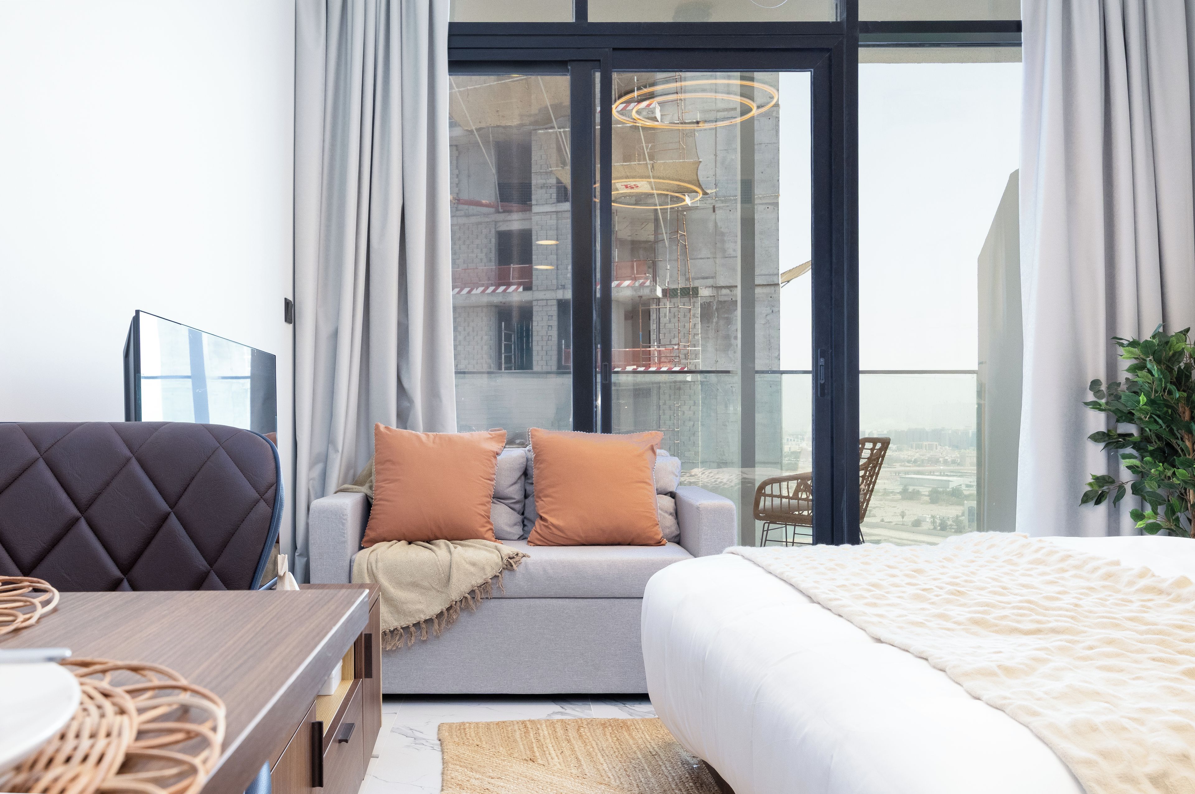 A modern bedroom with a sofa, a bed, a dining table, and a view of a cityscape through large windows.