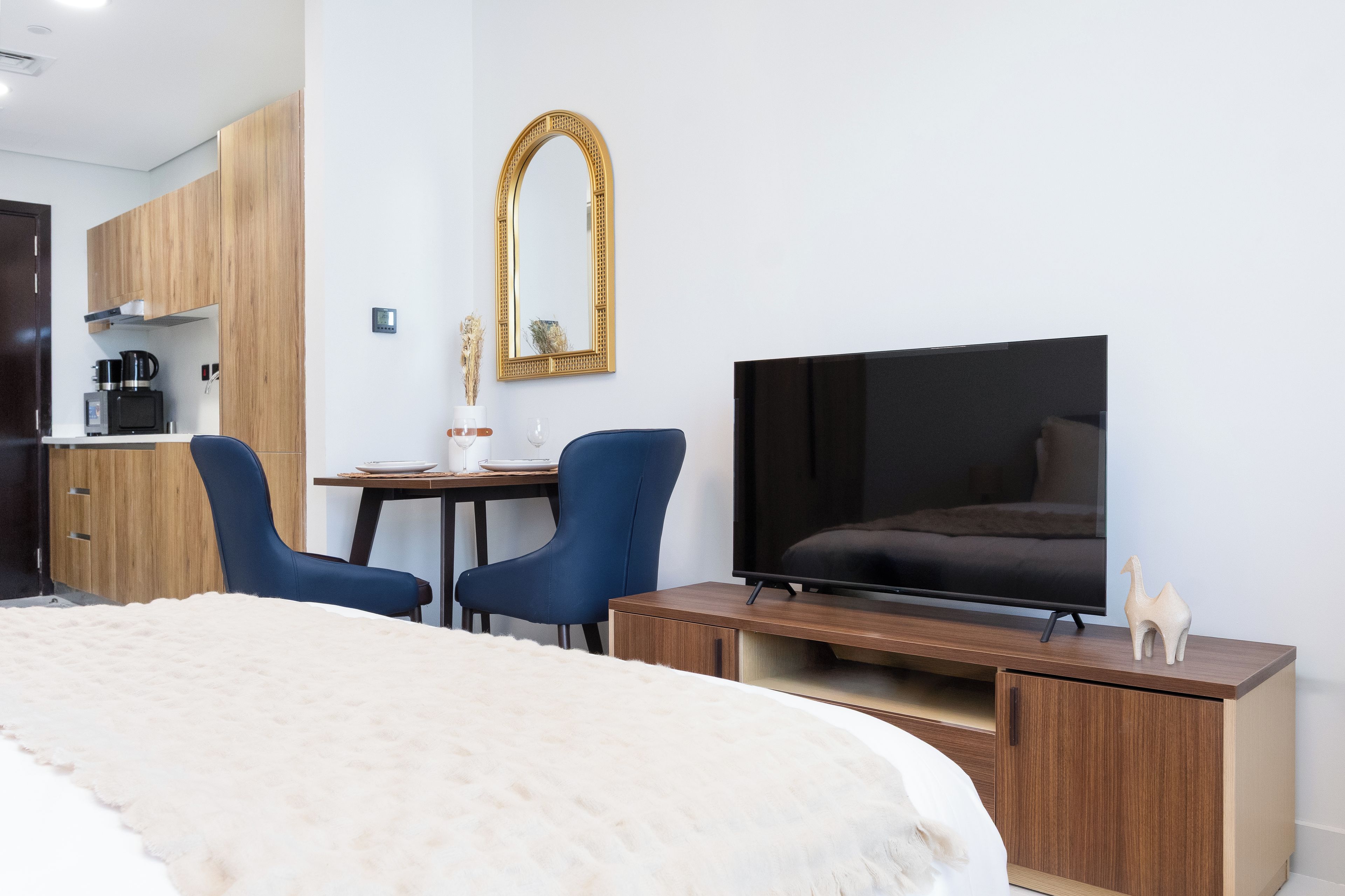 A hotel room with a bed, a TV, a desk with two chairs, a mirror, and a kitchenette.
