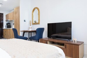 Et hotelværelse med seng, fjernsyn, skrivebord med to stole, spejl og et tekøkken.
