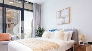 Een moderne slaapkamer met een groot bed, een nachtkastje met een lamp, een wanddecoratie en een uitzicht over de stad door grote ramen.