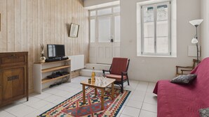 Una sala de estar acogedora con paredes de madera, un sillón rojo, una mesa de madera con una botella y vasos, un televisor en un estante y una ventana con vistas.