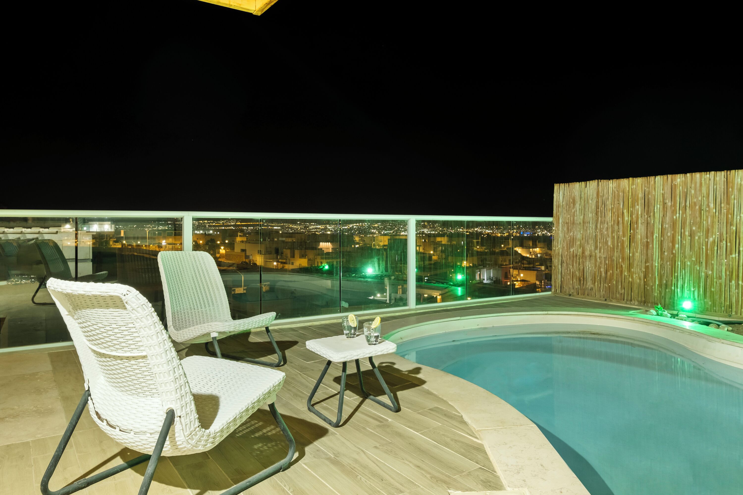 Área de piscina no terraço, com duas cadeiras de vime e uma pequena mesa, com vista para a cidade à noite.