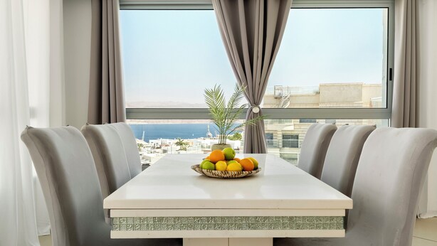 Ein Essbereich mit Tisch für zwei Personen, einer Obstschale und Blick auf Stadt und Meer durch große Fenster.