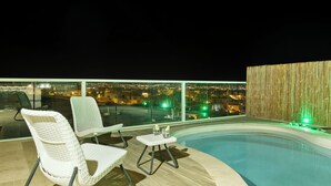 Ein Poolbereich auf dem Dach mit zwei Korbstühlen und einem kleinen Tisch, mit Blick auf eine nächtliche Stadtlandschaft.